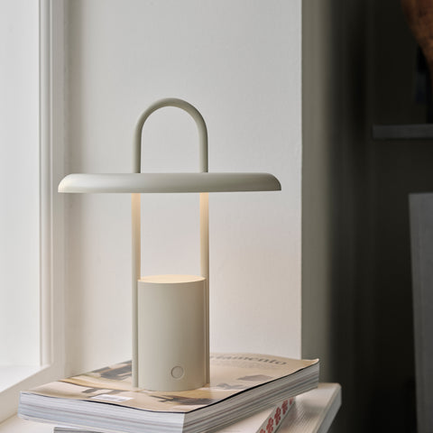 Sitting on a stack of magazines one a windowsill, an ecru-colored lamp with a cylindrical body, and a tall arched handle intersected by a wide shade emitting a downward glow.